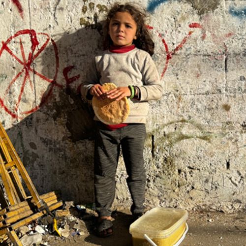 Et barn står intill en murvegg, ser i kamera og holder et brød i hånden.